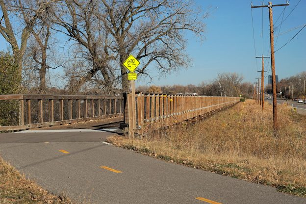 An asphalt bike and pedestrian path leads up to a wooden boardwalk, with a marshy area in the background.